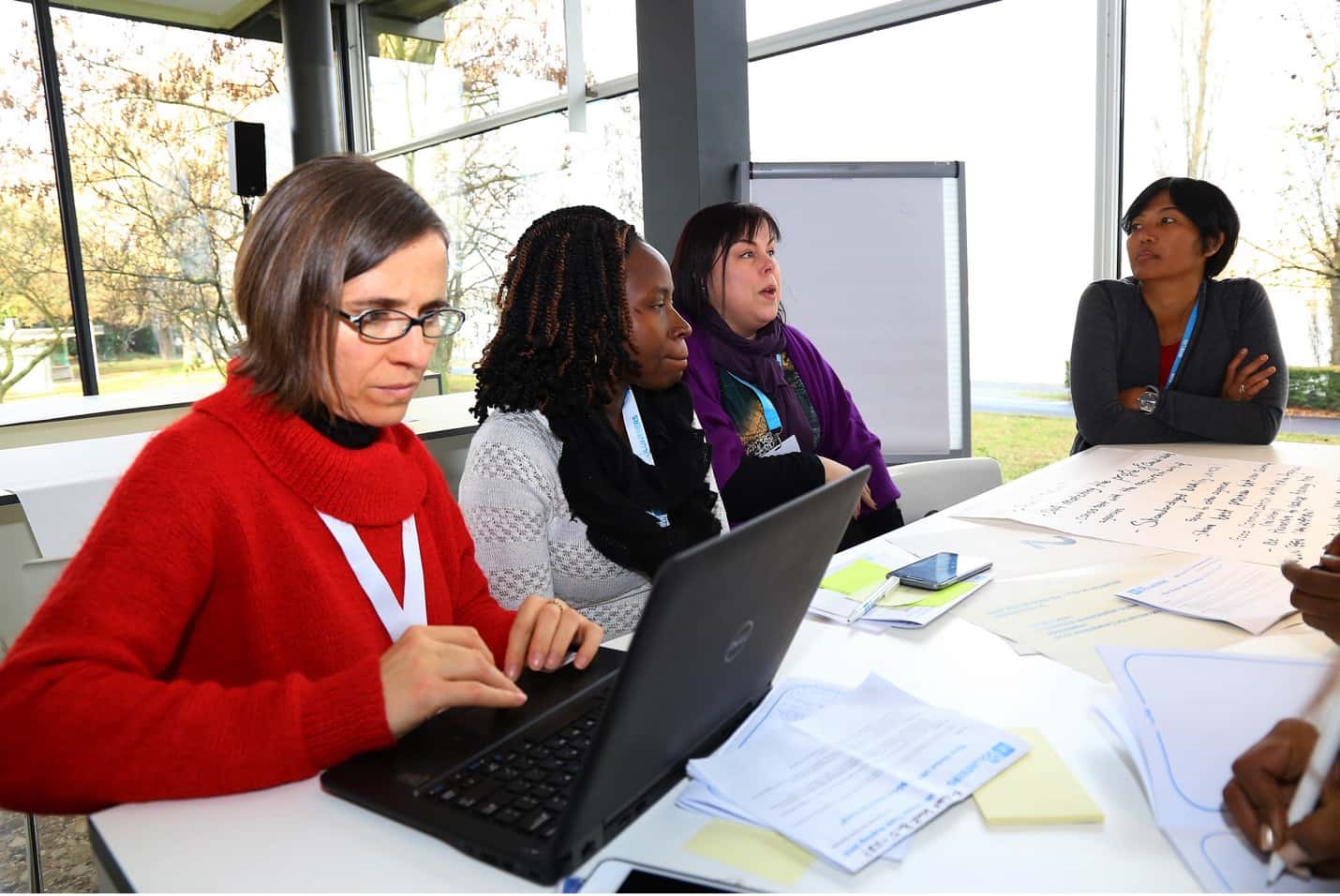 Four women at table, one of whom is working on her computer while other three converse. At workshop obtaining guidance and training on secure coding with industry-standard best practices, processes, and procedures.