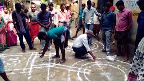 Men and women stand and watch as two men use chalk to display design on the ground. 