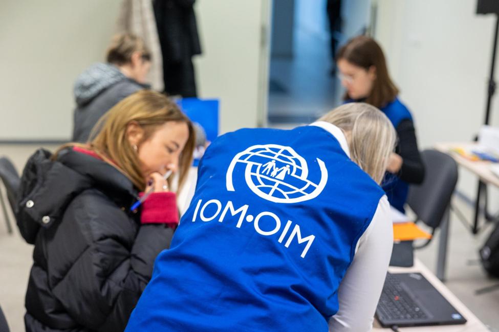 A staff member wearing a blue vest with the IOM–OIM logo assists a woman at a desk. In the background, other people are engaged in similar support activities.