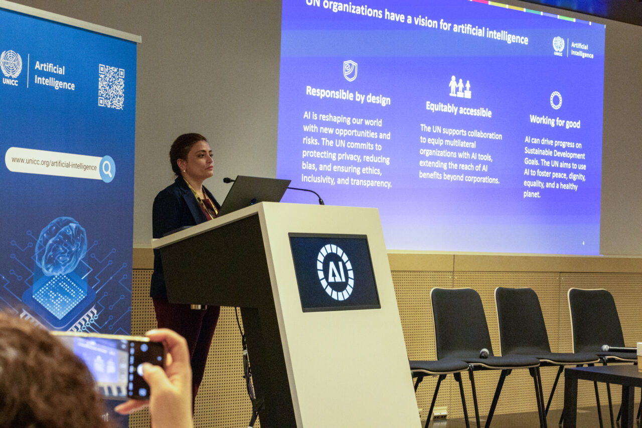 UNICC AI Hub Chief stands at a podium, presenting with a large digital screen behind her displaying key points about AI from the UN perspective. 