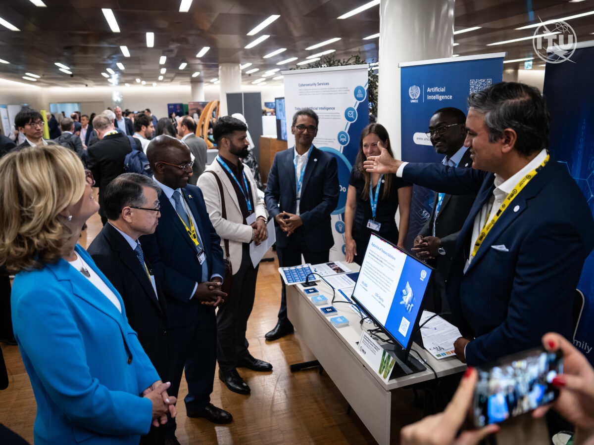 A group of delegates gathers around the UNICC booth at the exhibition during WSIS Week. UNICC Director in a blue suit is actively presenting information about UNICC's Artificial Intelligence services, gesturing toward a screen displaying key details. The booth features banners, brochures, and a monitor with a QR code.