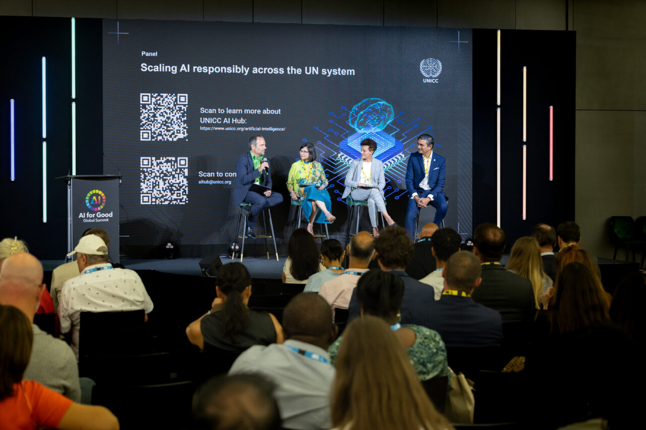 Four panelists, three seated and one speaking into a microphone, are on stage in front of a large screen displaying the UNICC logo, a digital brain graphic, and QR codes linking to more information about the UNICC AI Hub. The audience is seated and attentively watching the session. The setup includes a podium with the AI for Good logo and vertical neon lights as part of the stage design.