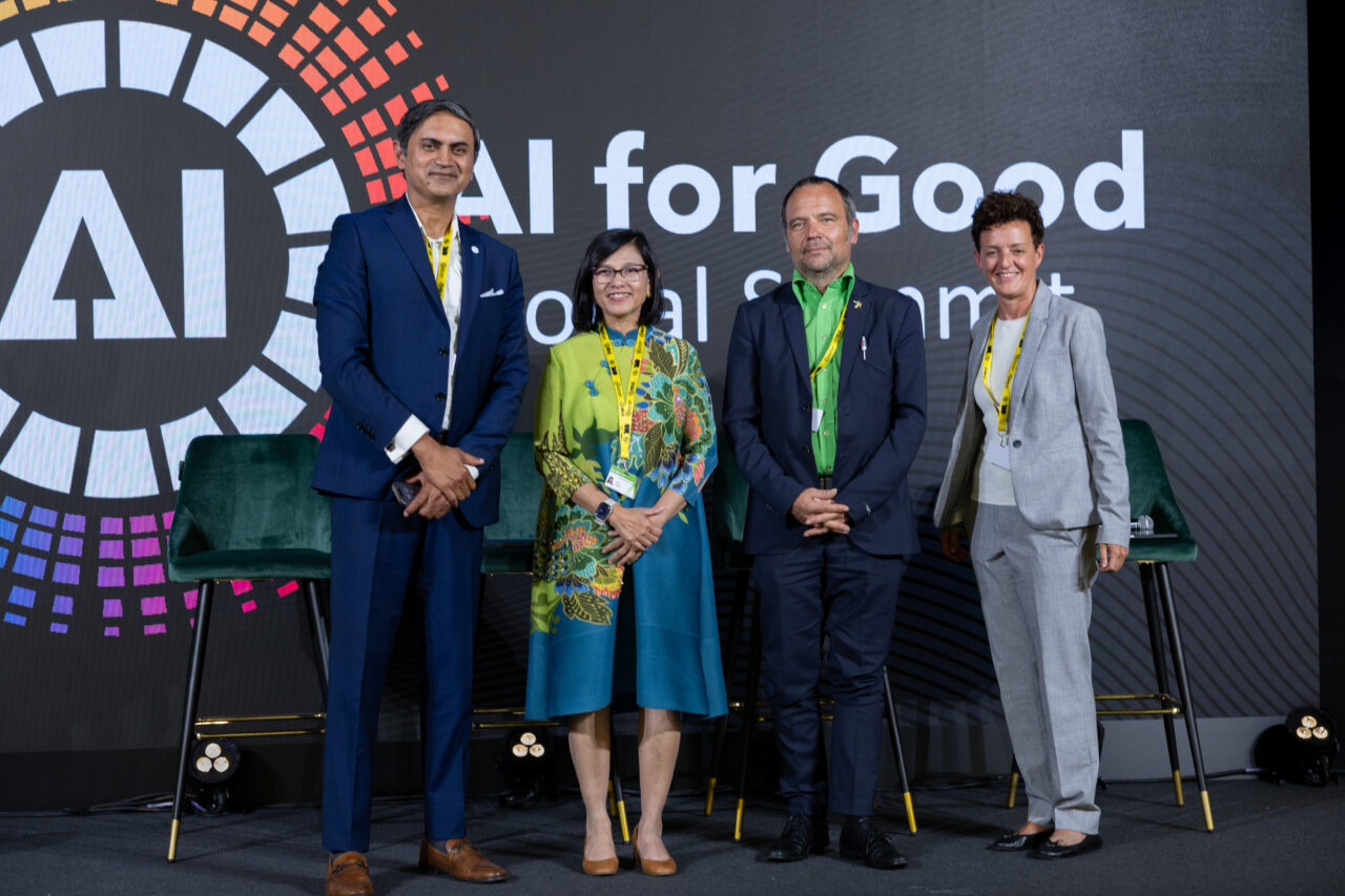 Four panelists stand on stage at the AI for Good Global Summit in front of a large screen displaying the summit's colorful logo and the words “AI for Good Global Summit.”