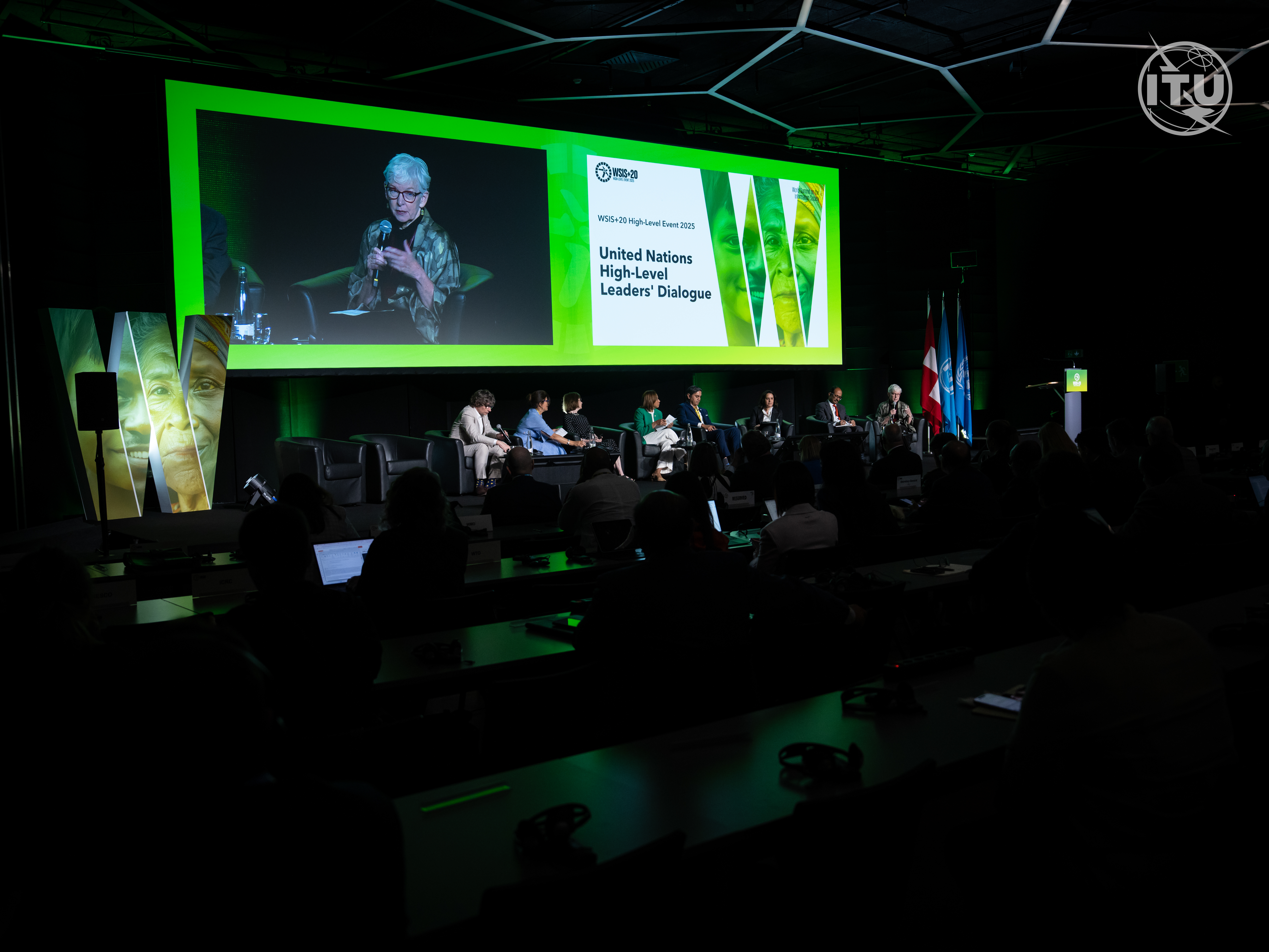 A wide view of a conference hall during the United Nations High-Level Leaders' Dialogue at WSIS+20. A panel of speakers is seated on stage under a large green-lit screen displaying the event title. The audience is seated in rows, watching the discussion.