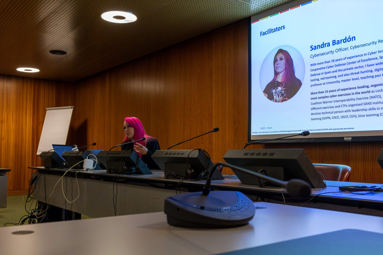 Speaker presenting during a UNICC session at Geneva Cyber Week, seated in a conference room beside a presentation slide introducing the facilitator.