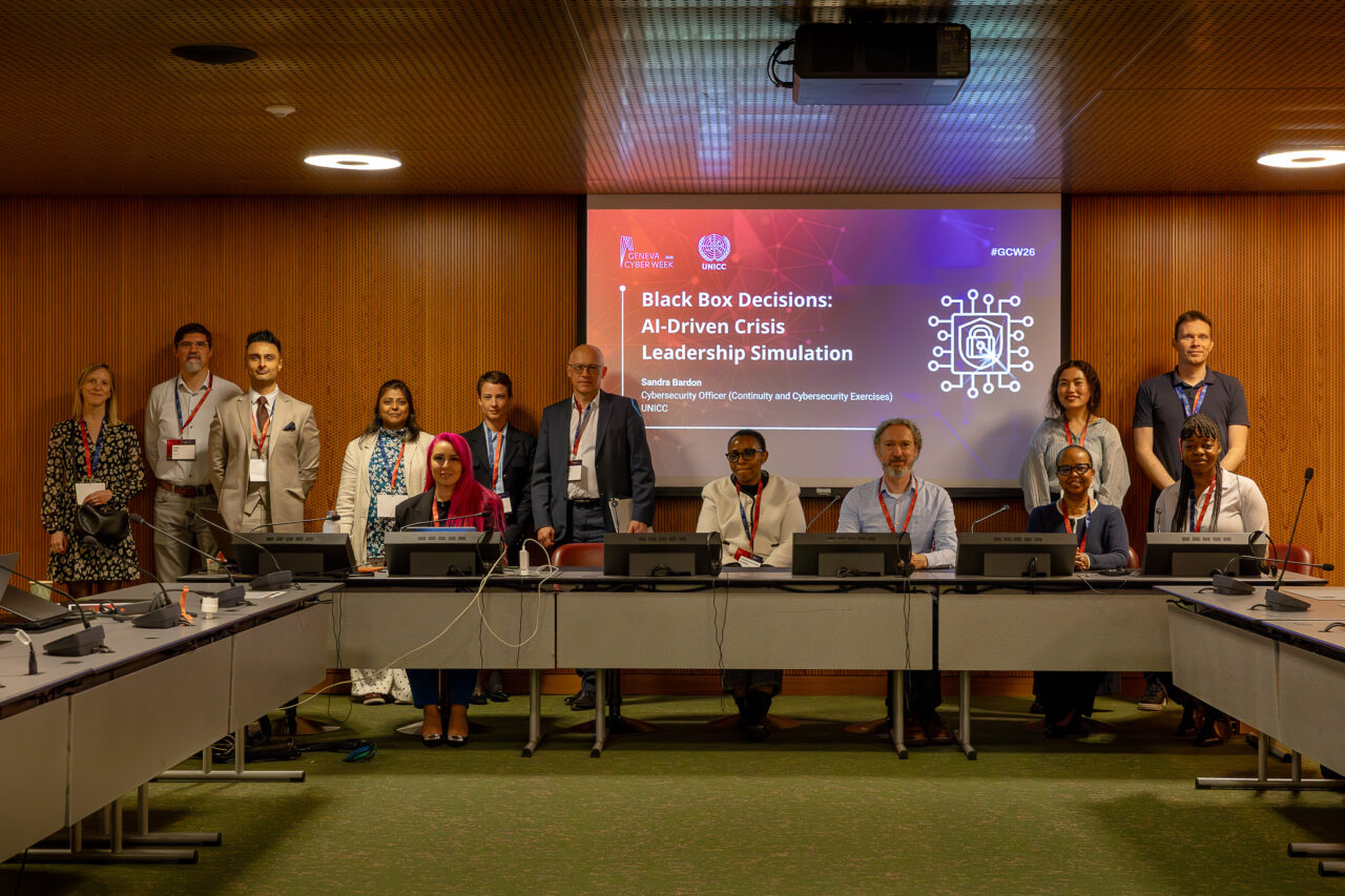 Group photo of participants at a Geneva Cyber Week session hosted by UNICC, standing and seated in a conference room in front of a presentation screen displaying “Black Box Decisions: AI-Driven Crisis Leadership Simulation.” The group includes speakers and attendees wearing conference badges, seated around a U-shaped meeting setup with microphones and monitors.